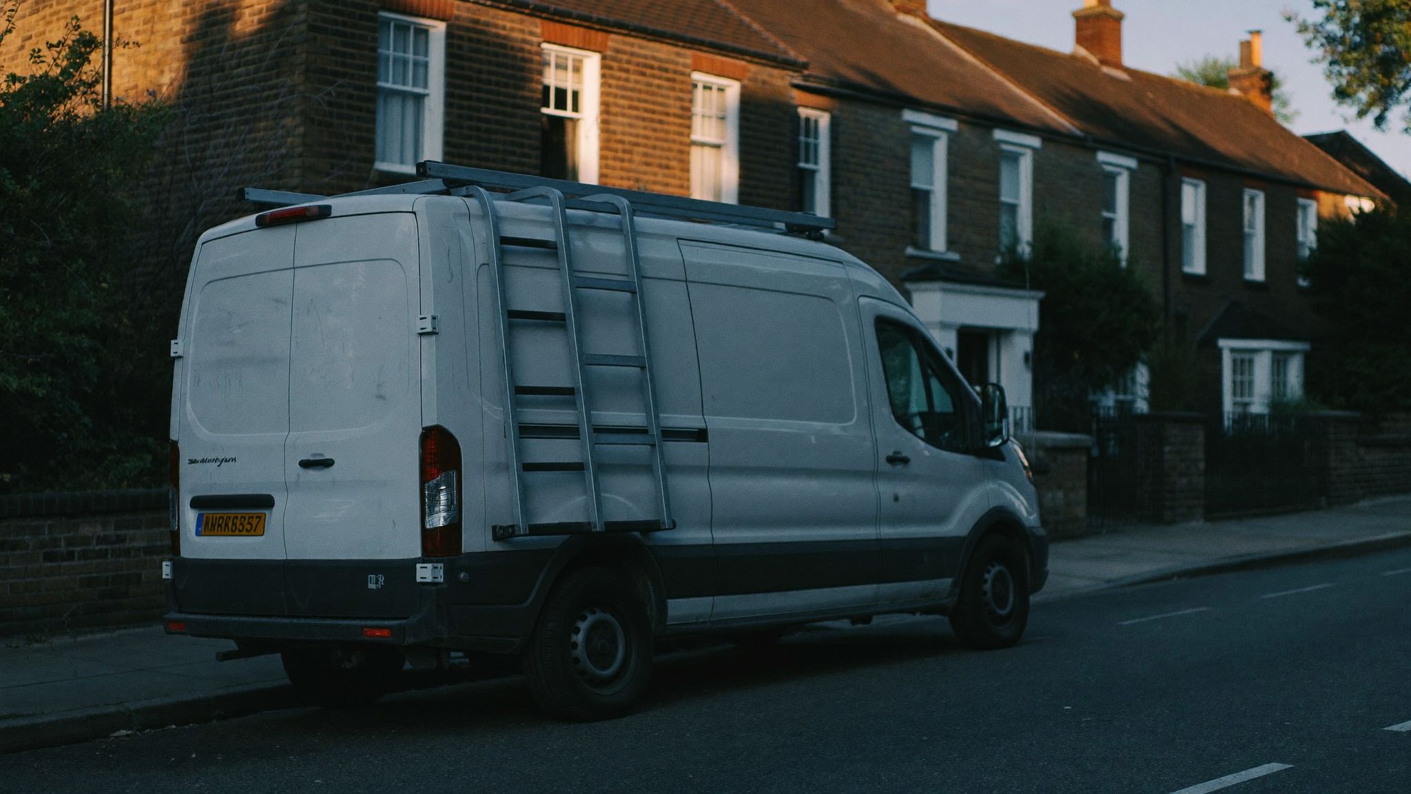 Unmarked white work van with a roof ladder outside a terraced house at golden hour